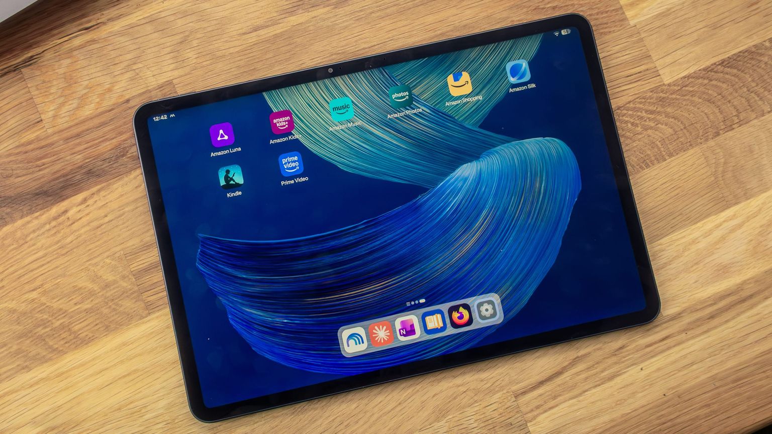 Alexa Tablet 12 Pro on a wooden table, home screen with the preinstalled Amazon apps and a dock with six apps